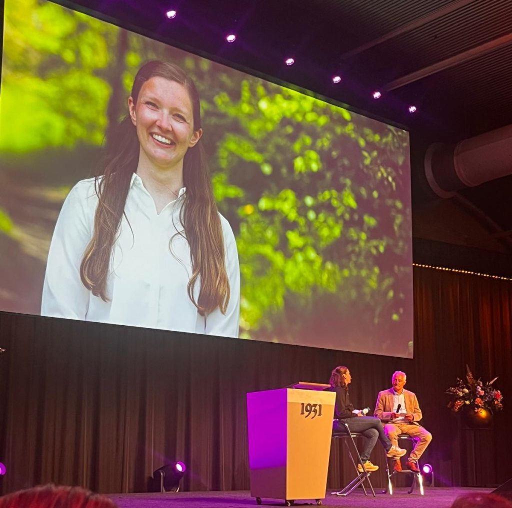 beeld van een evenement met een presentatiescherm waar een vrouw (lotgenoot SJK) op staat